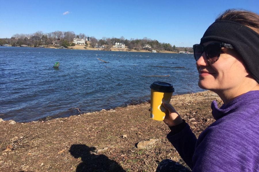 a woman wearing sunglasses holds a yellow coffee cup on the banks of a river on a clear and sunny day