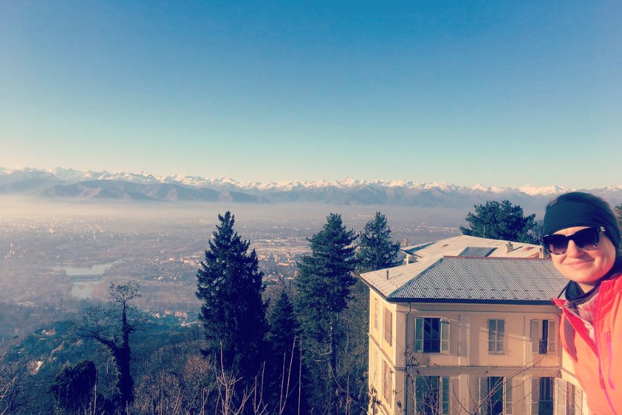 A woman in a hat and shades being photographed inf ront of the snowy alps and the city of Turin far below.