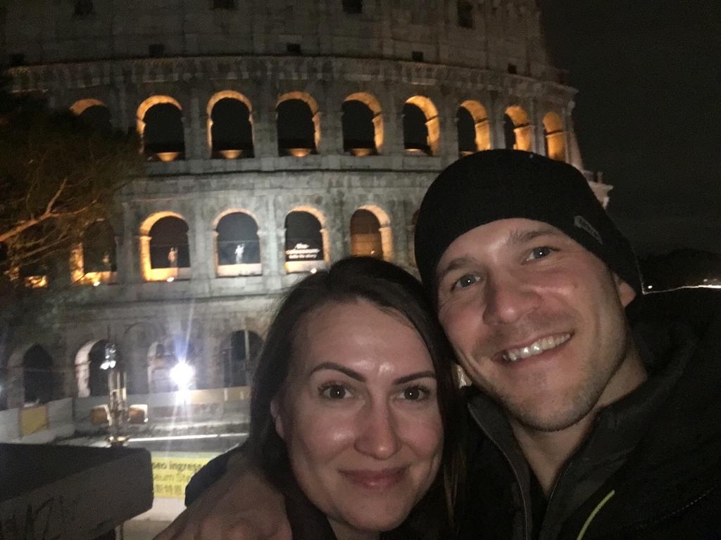 a man and woman smile for a selfie in-front of the Colleseum in Rome at night.,