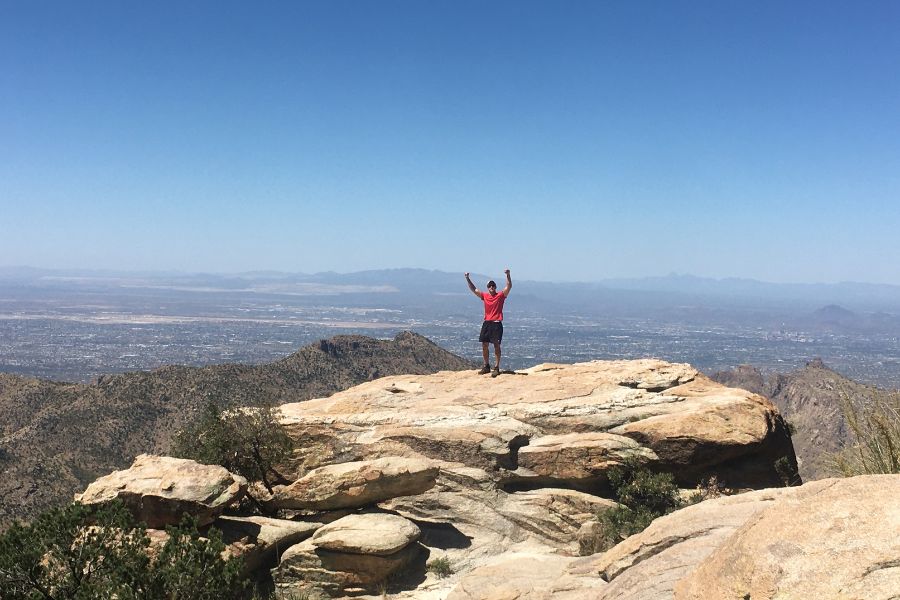 A man raises his hands over his hand high up on a mountain top that overlooks Tucson, AZ, thousnads of feet below. 