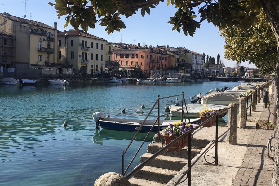 The banks of Lake Garda, with boats moored and sunshine hitting the colorful buildings.