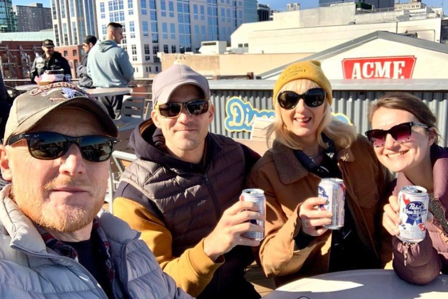 4 adults, two men and two women sit on a rooftop bar on a sunny day drinking PBR cans of beer.