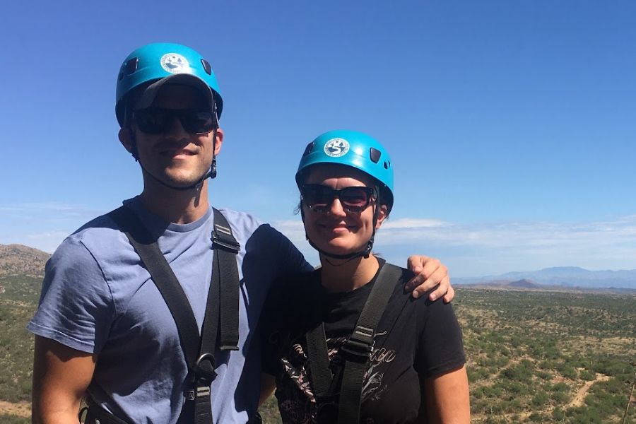 man and a woman wearing helmets and harnesses standing in a desert scene ready to zipline. 