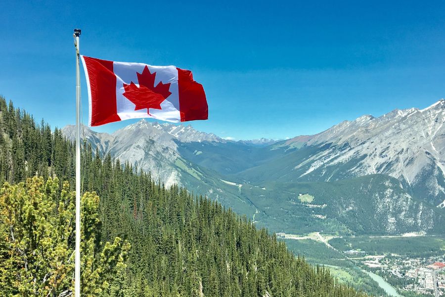 The Canadian flag flies high and proud on hills of green trees and snow covered peaks in the distance.