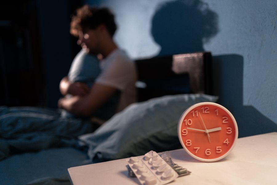 a man lies awake and hugs a pillow while a red clock shows it to be 2.45 am. Sleeping pills sit next to the clock.