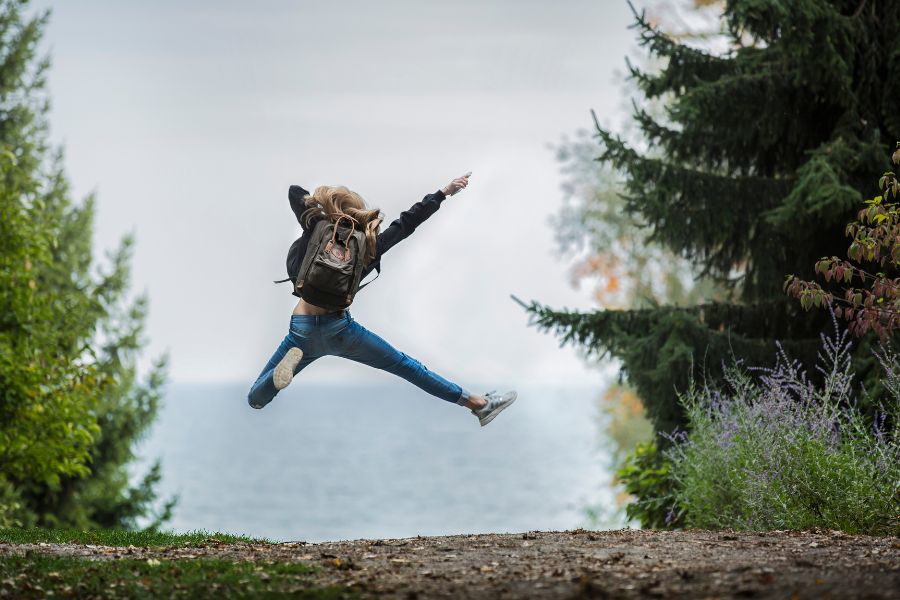 A girl with a rucksack leaping into a star in the air whilst hiking on a path towards the ocean