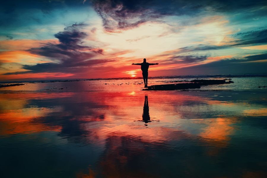 a man stands on a rock in still water as the sun sets. His arms are spread open wide