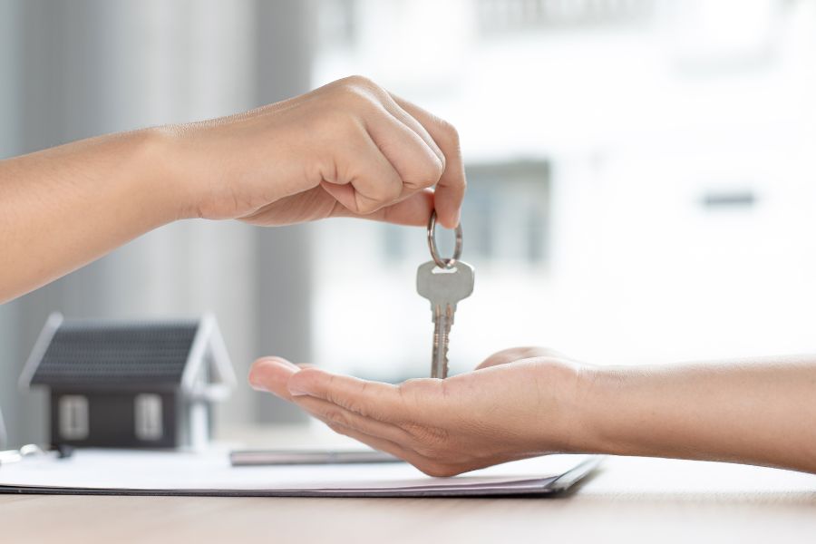 one hand passes a single silver key to another hand. A small toy house sits in the background