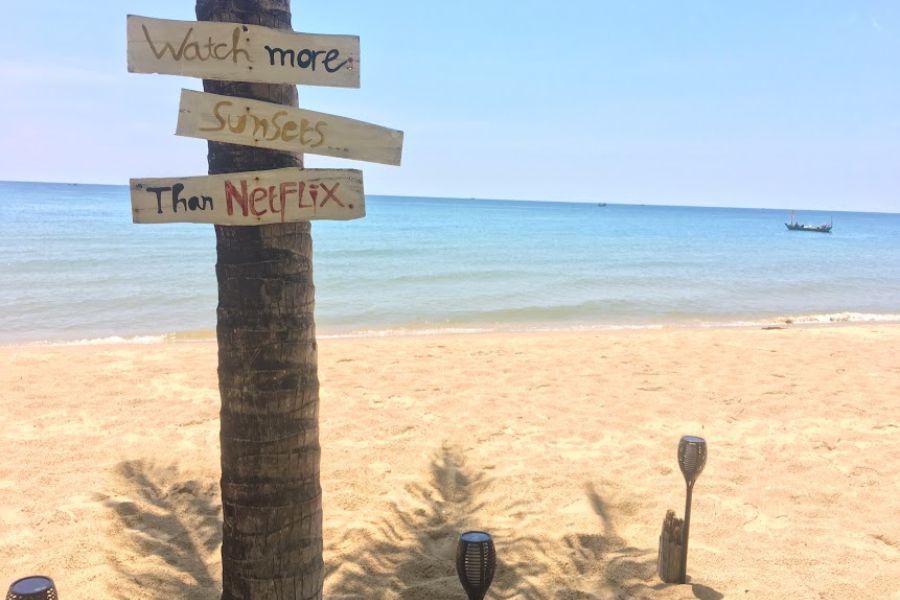 a stunning beach seen, blue water, white sands and a hand painted sign on a tree that read 'watch more sunsets than Netflix'