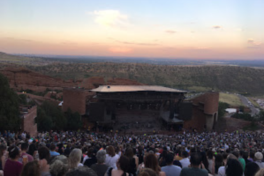 a packed seated audience looks ahead towards a band playing on the Red Rocks stage. In this distance the sun sets