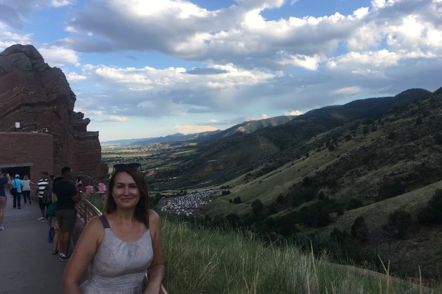 A woman poses for a selfie with the Red Rocks arena and a far stretching green and hilly landscape behind her