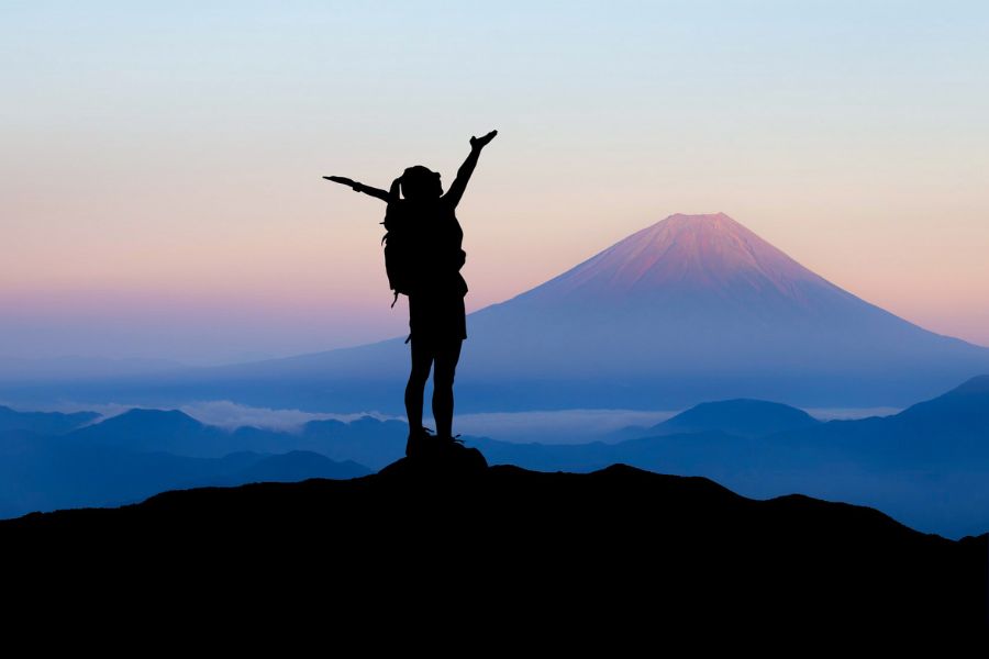 a backpacker stands a top of a hill arms raised looking over a volcano scene