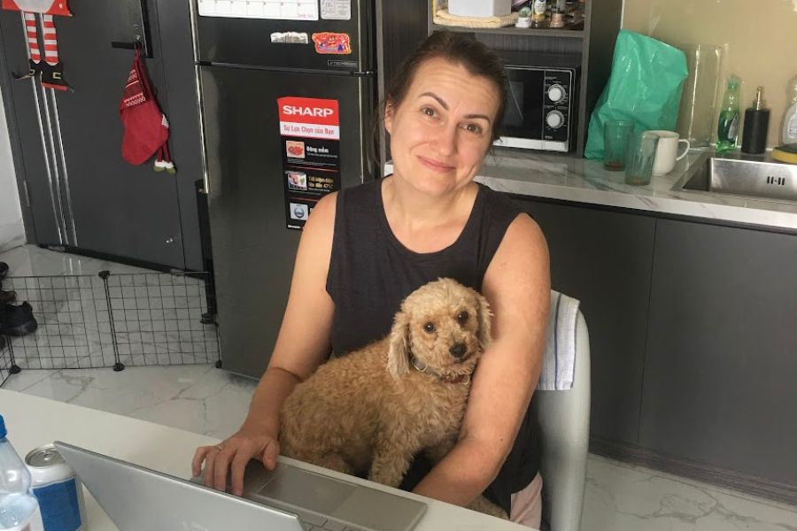 a woman sits working at a laptop with a dog sitting on her lap, both look at the camera