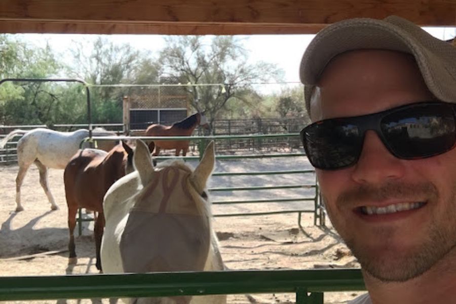 a man takes a selfie with 4 horses in a coral.