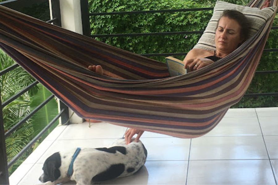 a woman lays in a hammock on a balcony in Costa Rica reading a booking with one hand and stroking a black and white dog on the floor with the other