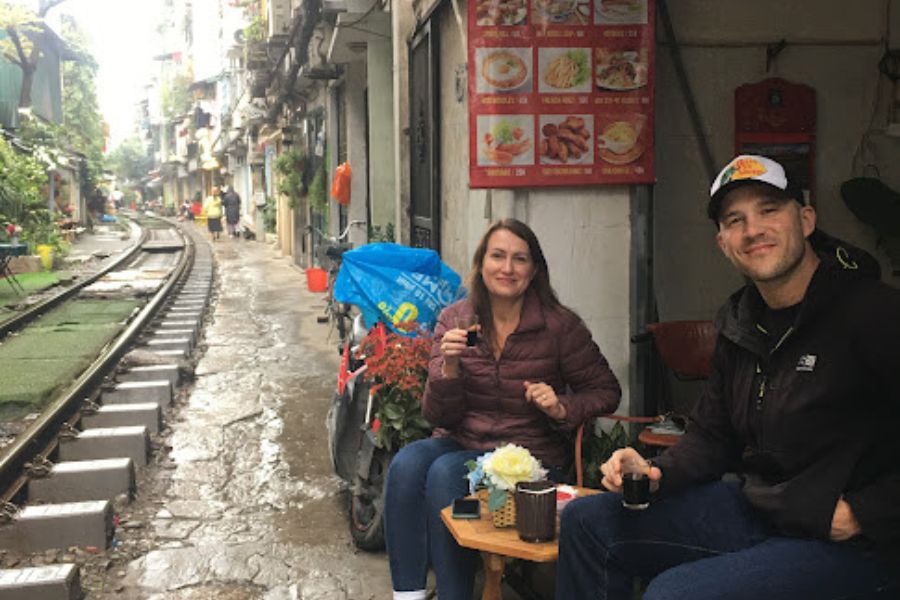 2 people sit close to a railway track drinkiong coffee